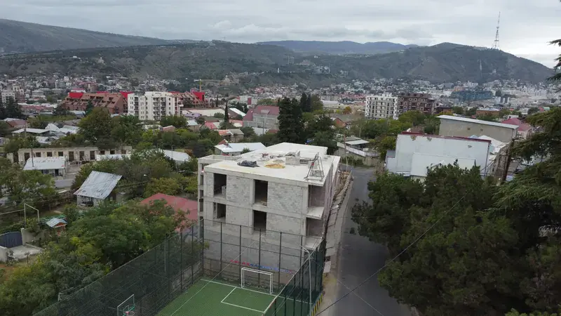 Aerial view of the four-level concrete shell of the Avlabari building with the Tbilisi cityscape and hills behind it and the surrounding residential neighbourhood