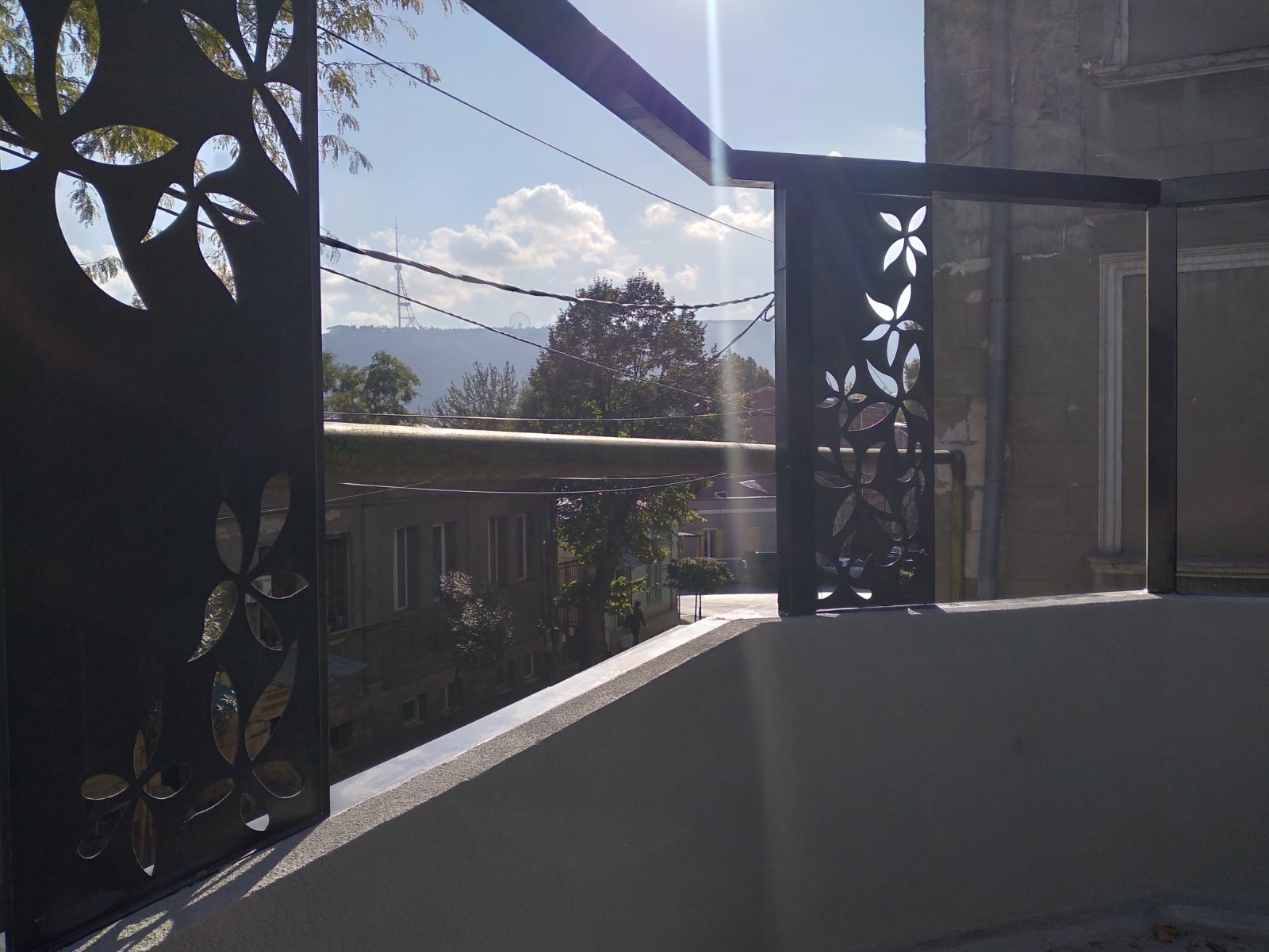 Rooftop terrace perspective showing decorative metal screens with floral patterns, views toward Tbilisi hills and historic neighborhood buildings